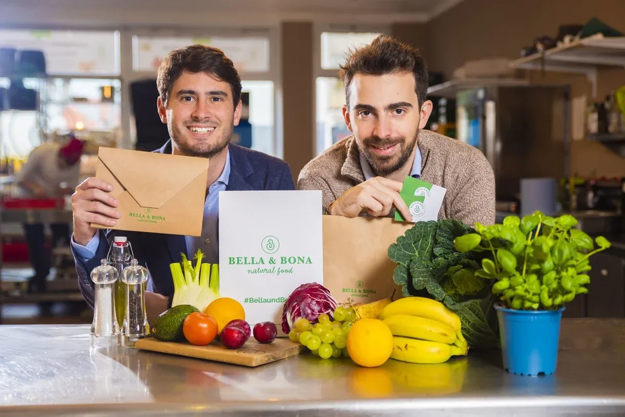 Founders of Bella&#x26;Bona surrounded by fresh food, highlighting their commitment to quality and sustainability.